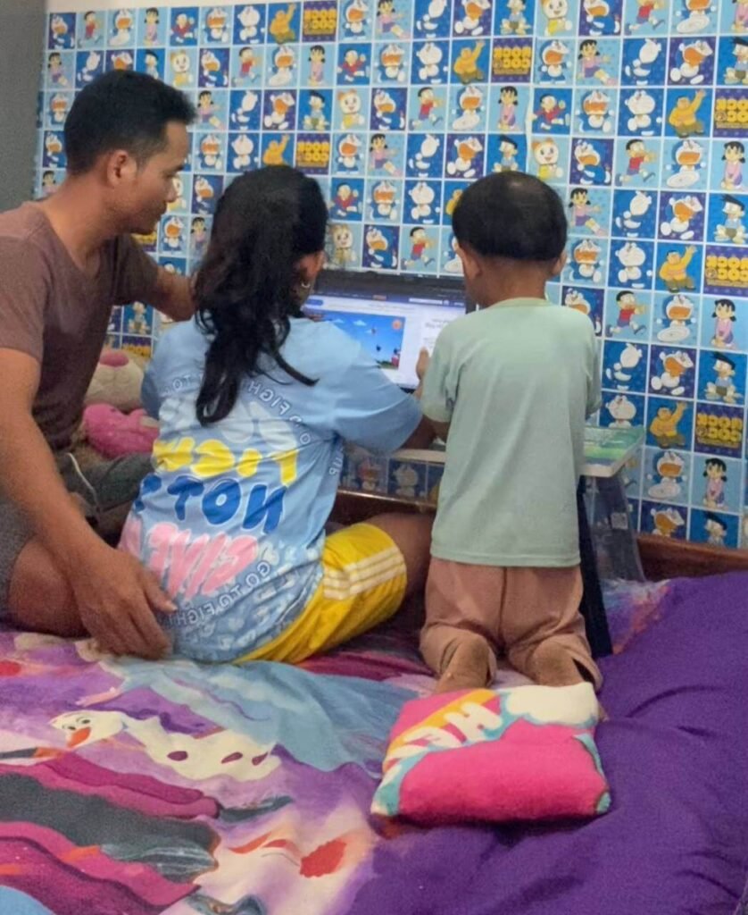 A father sitting on a bed with his daughter and younger son, patiently helping them look at a laptop screen together.