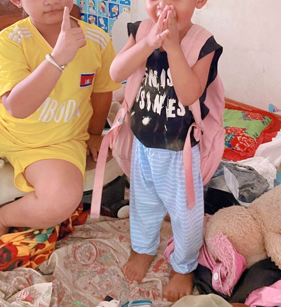 A young girl wearing a bright yellow Cambodia sports jersey standing next to her younger brother who is wearing a pink backpack.