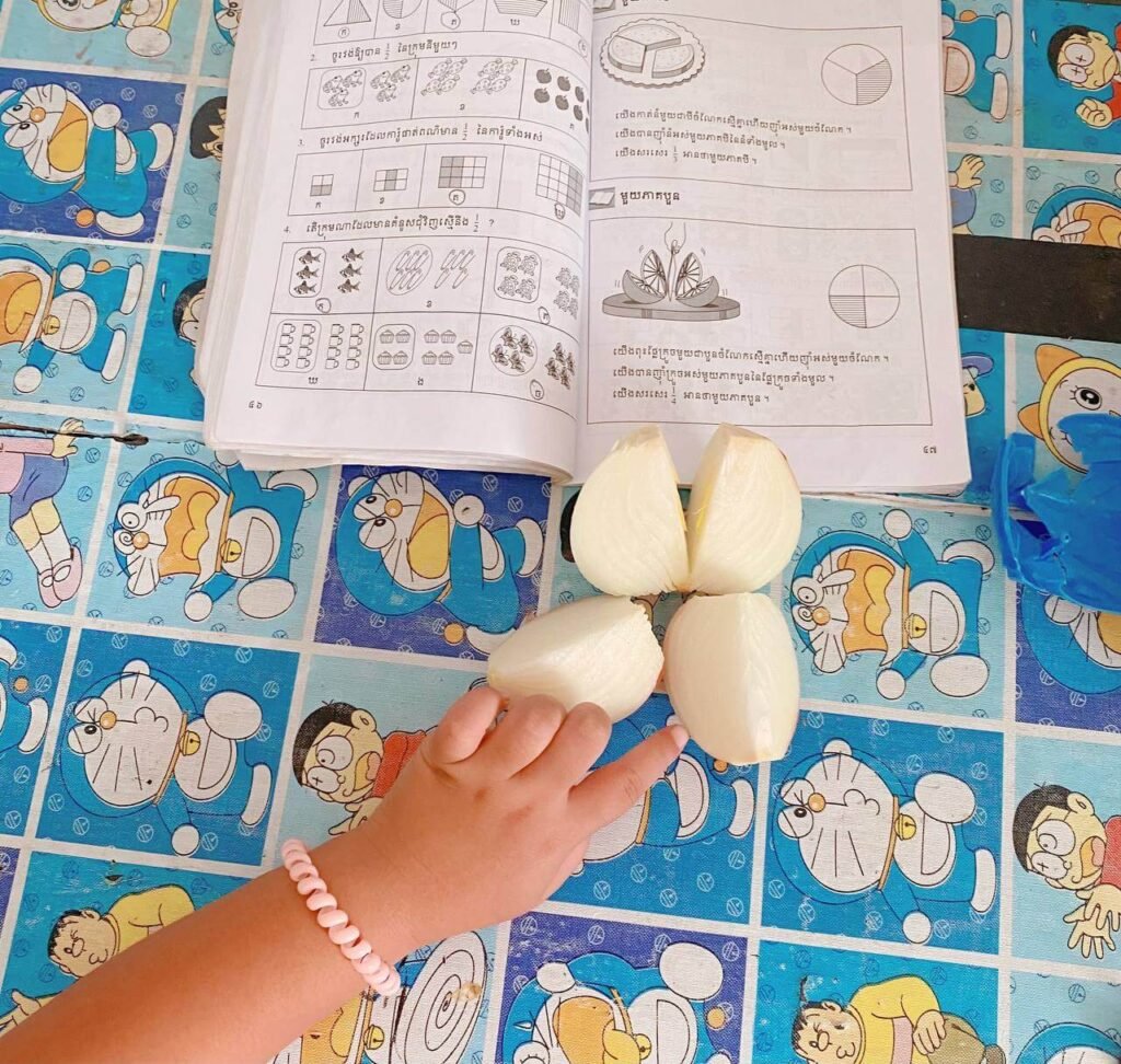 A top-down view of a textbook with fraction diagrams, next to a white onion cut into four distinct quarters, being pointed at by a child's hand with a pink bracelet.