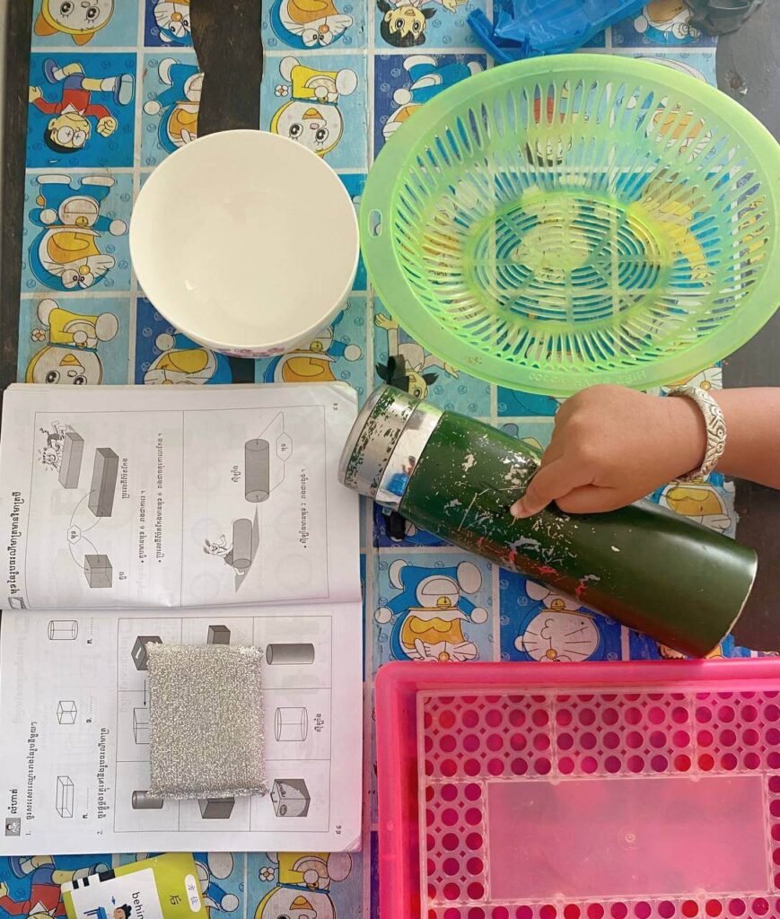A child's hand pointing to a green cylindrical water bottle, using household items like a white bowl and baskets to match the 3D shapes in their open math textbook.