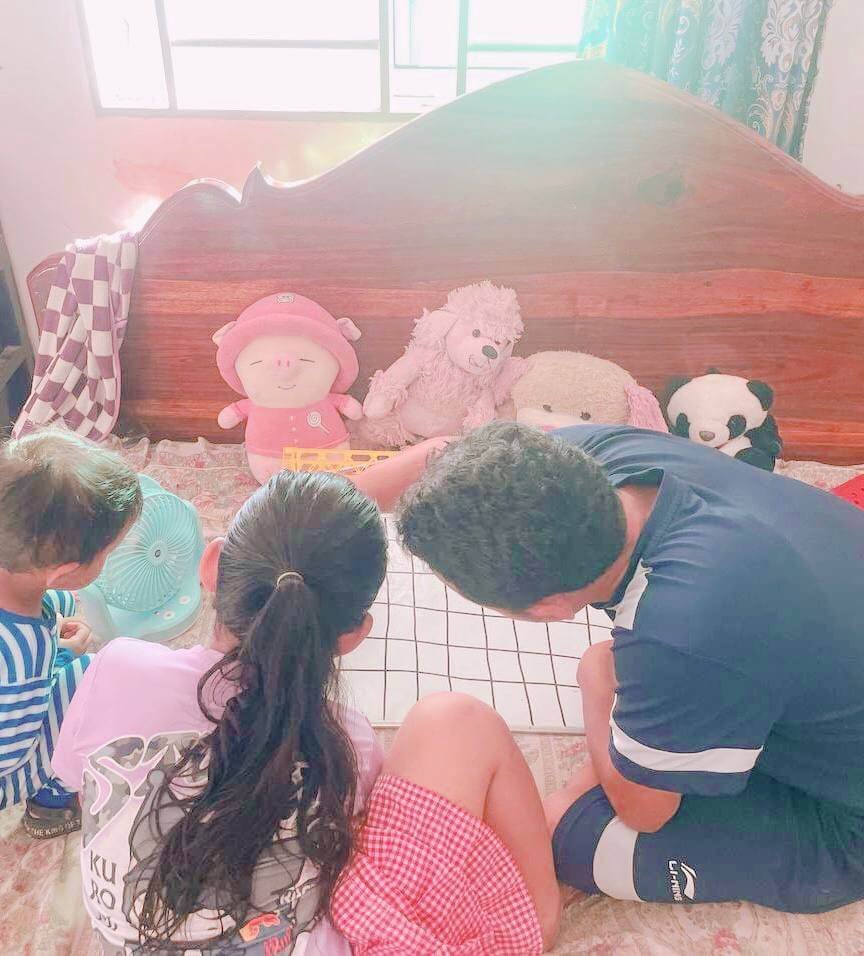 A father and two young children sitting on a bed together, looking over a blank whiteboard grid to map out a morning routine schedule.