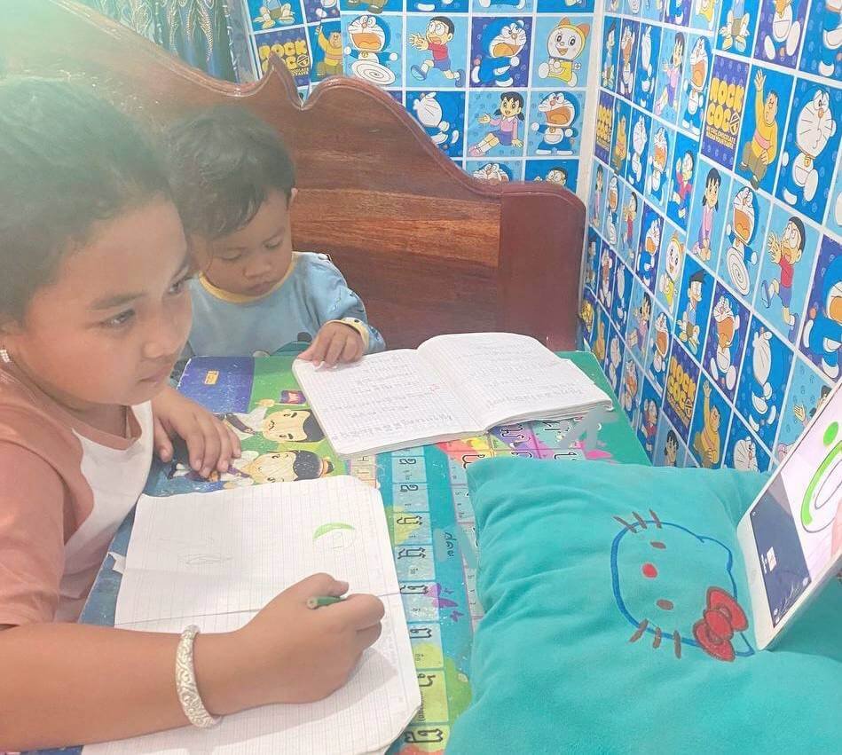 An older sister writes in her notebook while her younger brother sits beside her, quietly looking at his own book.