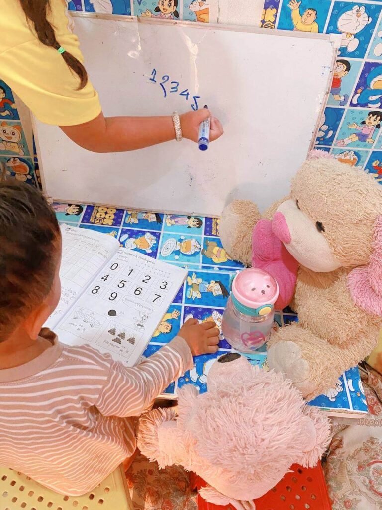 An older sister writing numbers on a whiteboard to teach her younger brother, who is looking at a counting workbook next to a teddy bear.