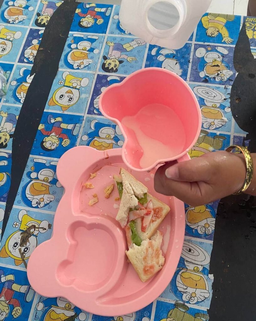 A child's hand holding a pink plastic cup over a plate with a sandwich, with milk accidentally spilled across the plate and table.