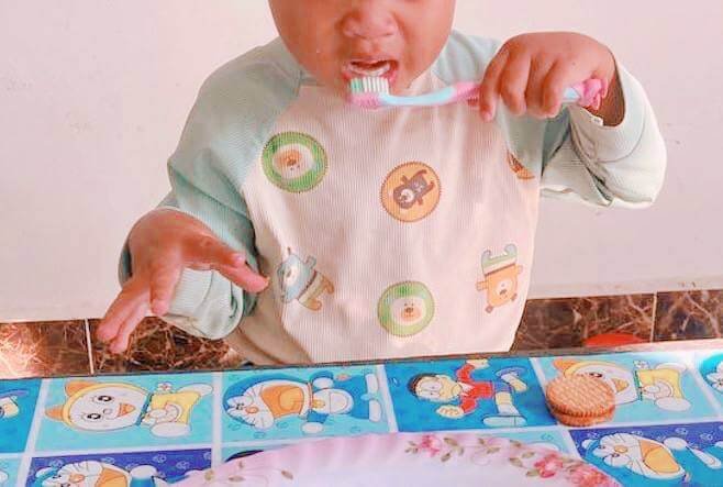 Close-up photograph of a toddler holding a toothbrush to his mouth, sitting at a table covered by a colorful Doraemon tablecloth with a biscuit nearby.