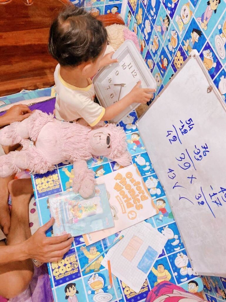 A toddler holding a white square clock over a study table with a phonics book, a pink stuffed dog, and a whiteboard.