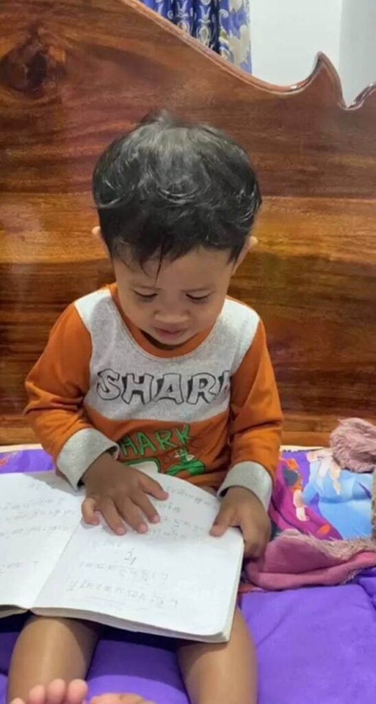 A toddler sitting on a bed independently flipping through an open notebook.