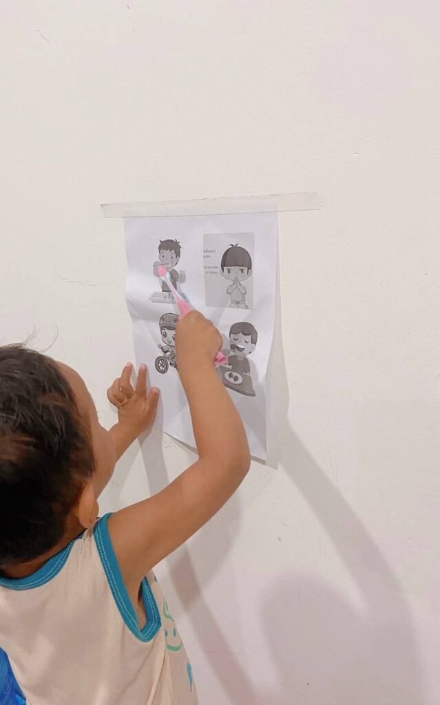 A toddler with dark hair, wearing a white tank top, stands in front of the visual chart on the wall. He is holding a pink toothbrush and pointing it directly at the illustration of a child brushing teeth.
