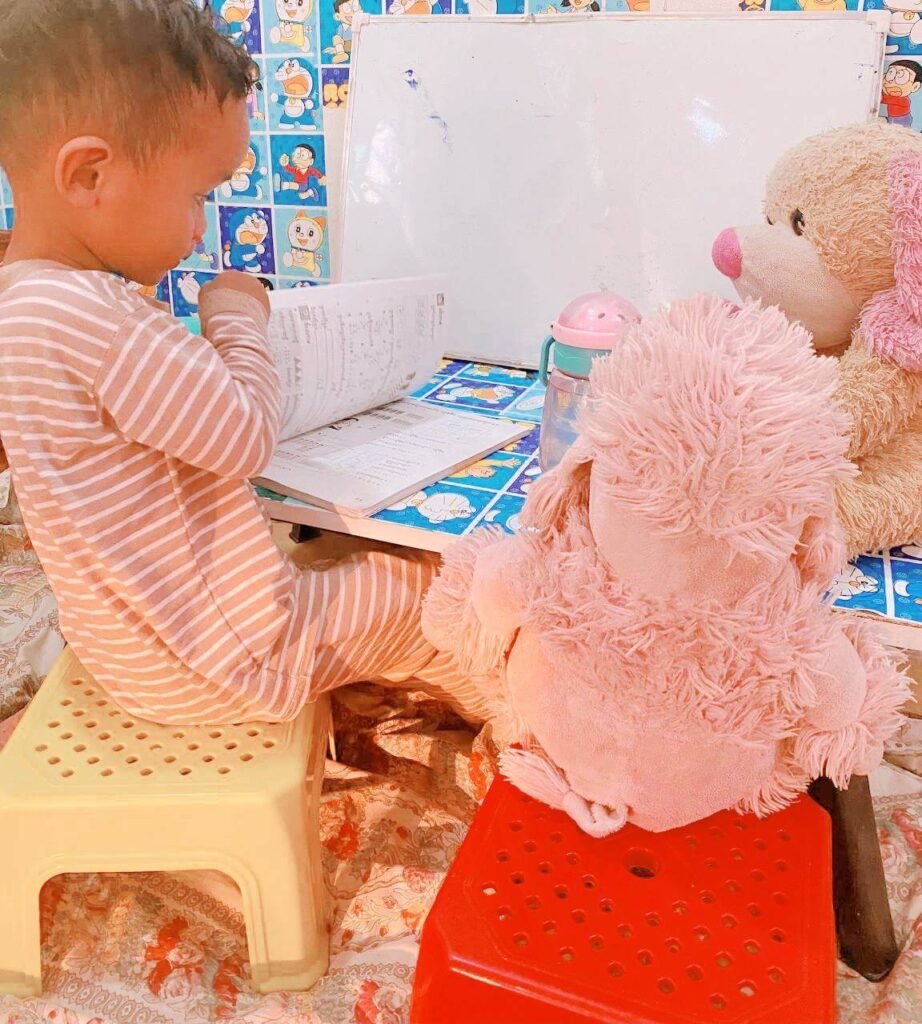A toddler sits on a yellow stool at a small table, looking at an open book with two stuffed dogs placed next to him.