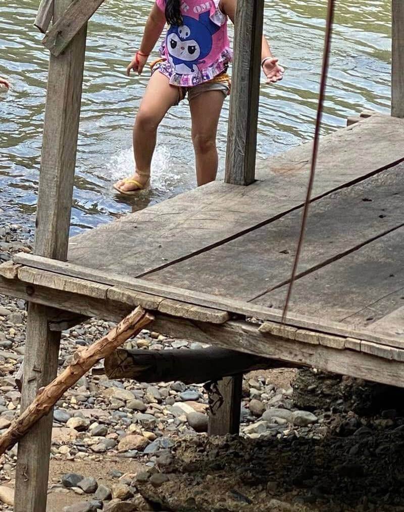 A young girl cautiously stepping off a rustic wooden dock into a shallow, rocky river.