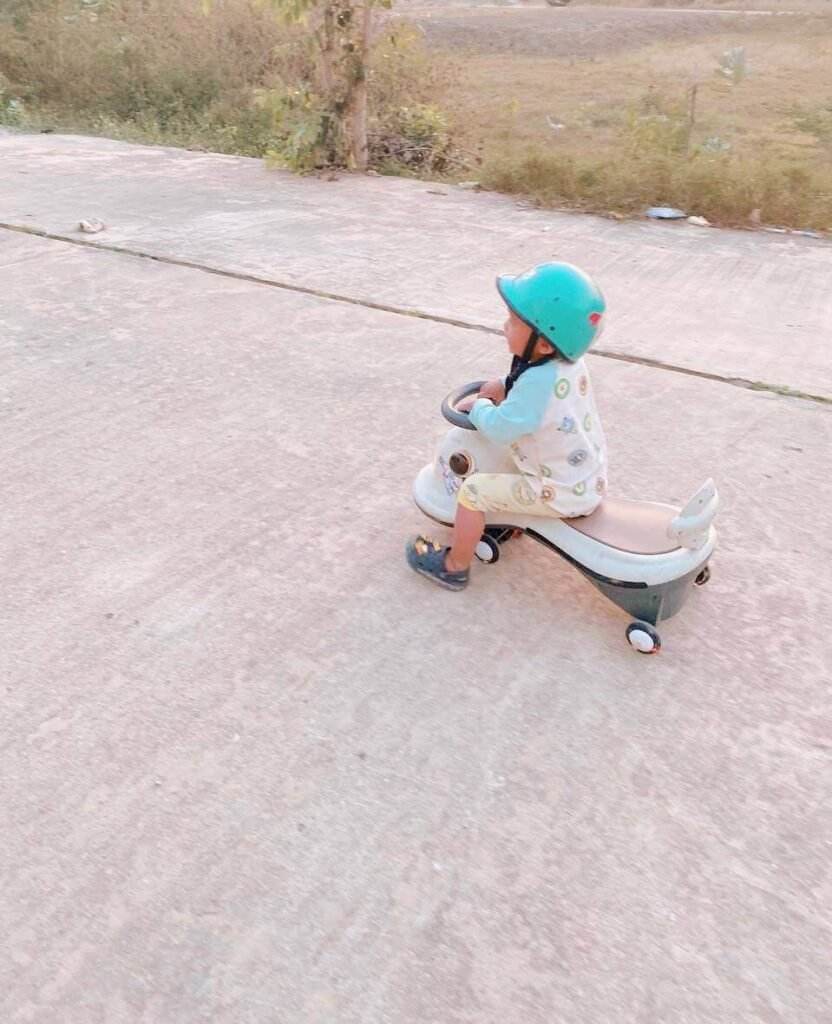 A child wearing a bright blue helmet sits on a white and grey ride-on toy on a concrete path outdoors at dusk, demonstrating a consolidated safety habit.