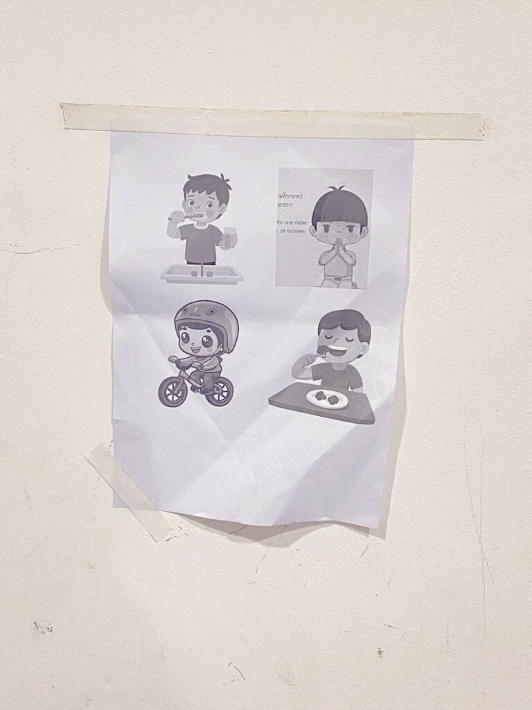 Close-up photograph of a printed visual routine chart taped to a white wall with masking tape, featuring four simple illustrations: brushing teeth, sampeah greeting, wearing a helmet on a bike, and eating.