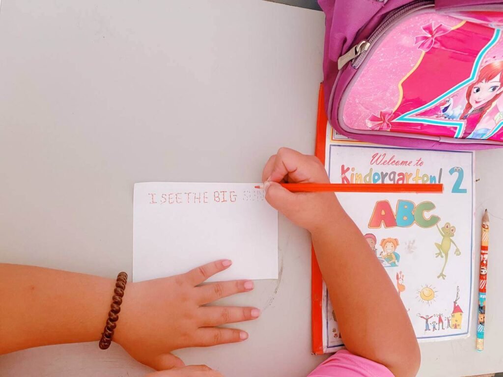 A child writing the simple sight word sentence 'I see the big sun' on a blank card next to their school materials.