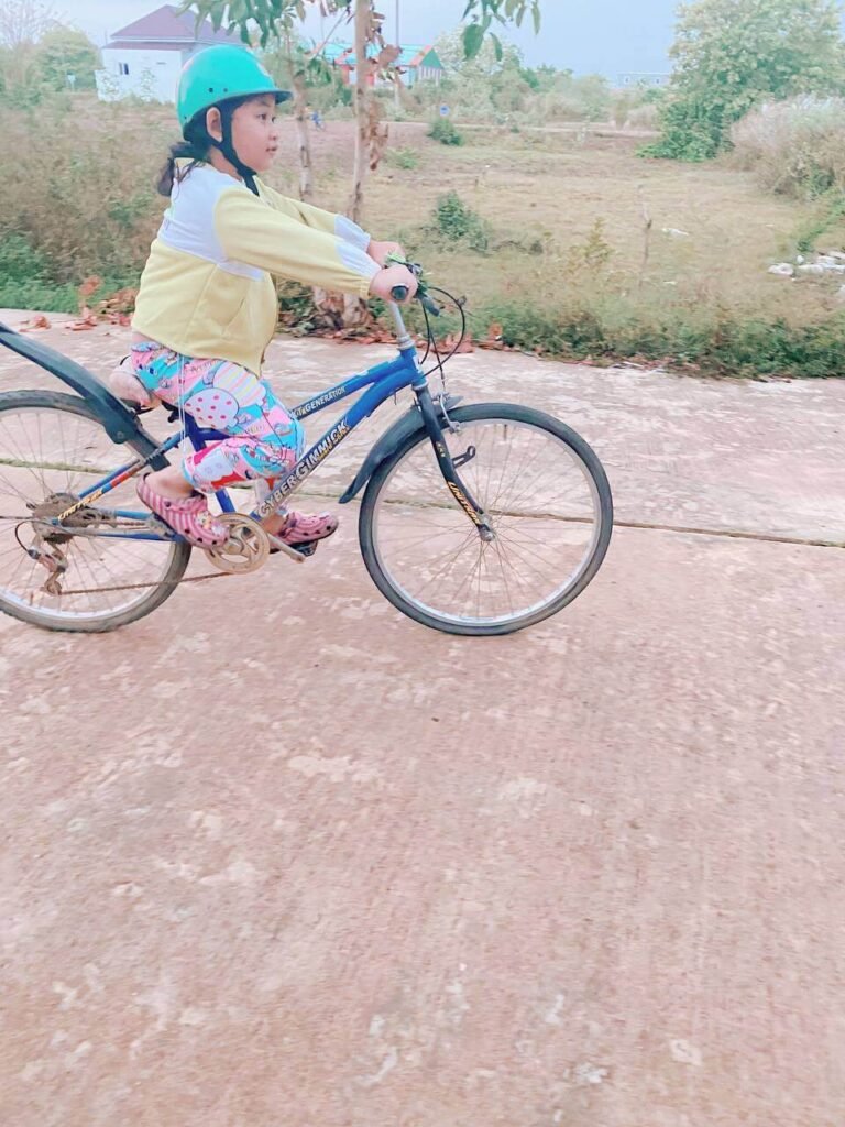 A young girl wearing a green safety helmet confidently riding a blue bicycle down a concrete path.