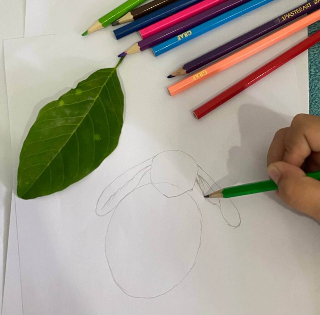 A real green leaf placed on white paper next to a child's pencil sketch of bunny ears, showing how the ears mimic the leaf's shape.