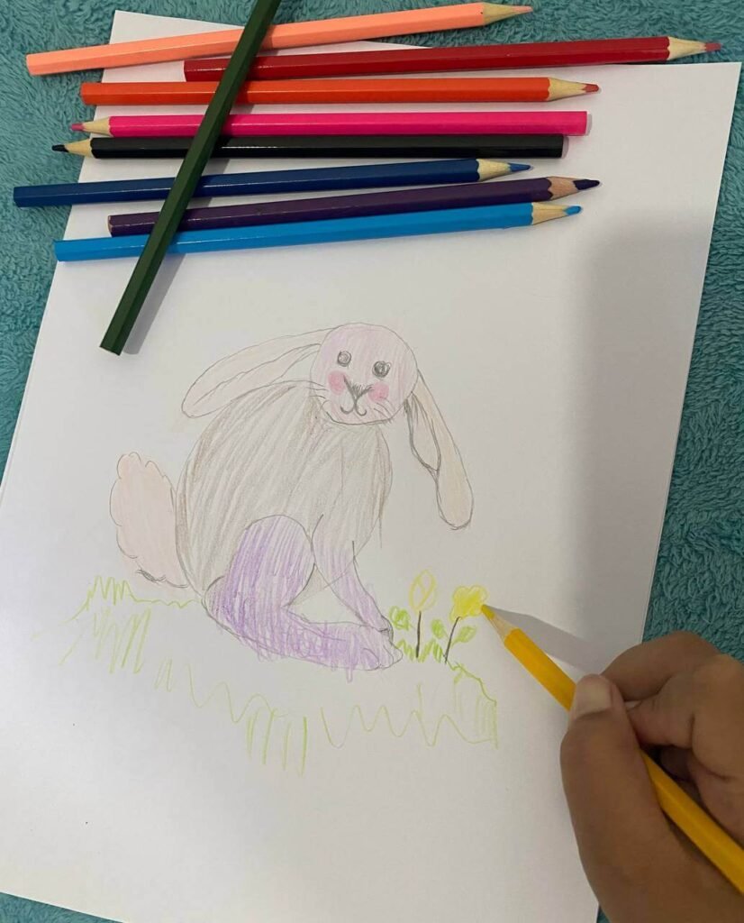 A child's hand coloring yellow flowers next to a finished drawing of a bunny with a pink face, gray body, and purple legs.