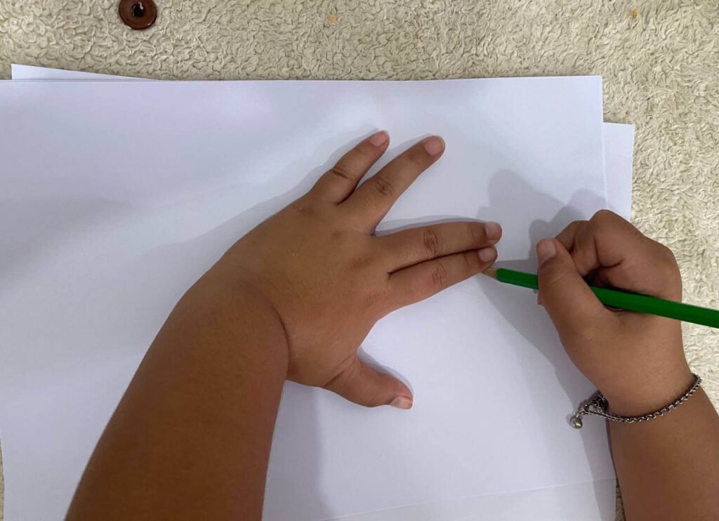 A top-down view of a child's left hand pressed flat against white paper with fingers slightly spread, while their right hand traces the outline with a green pencil.