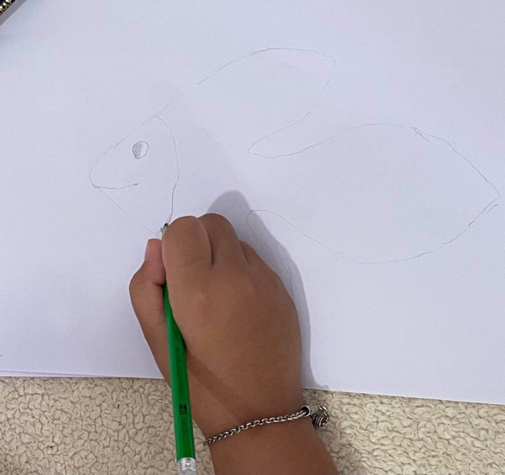 A child's hand using a pencil to draw a simple eye and mouth on a fish outline traced from a leaf.