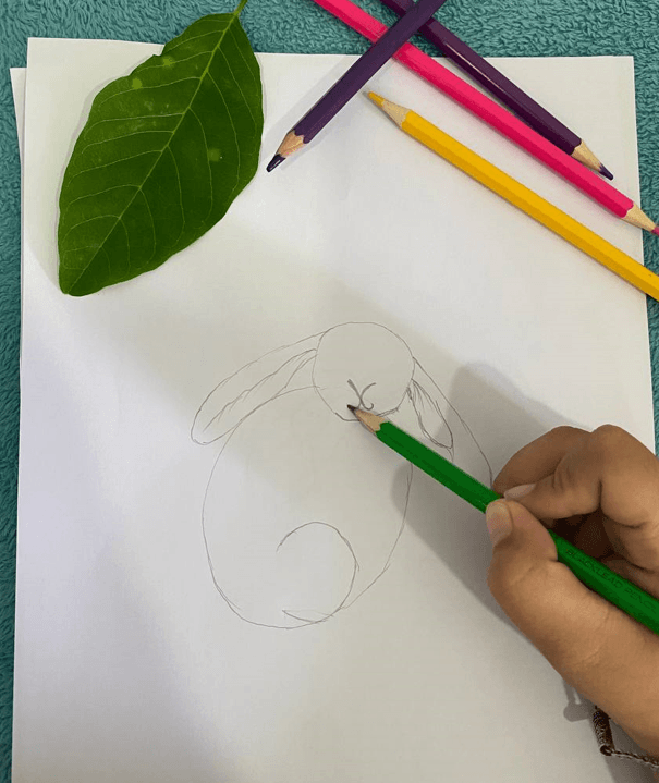 A child's hand using a green pencil to draw a Y-shaped nose and mouth on a sketch of a bunny.