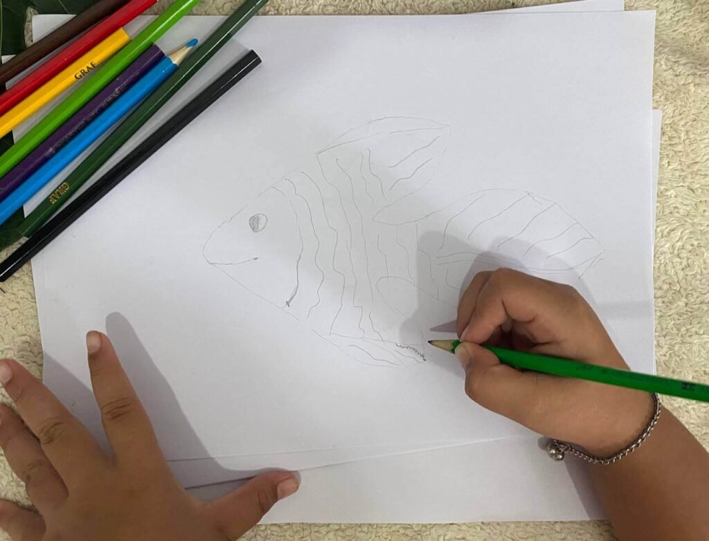 A child drawing wavy lines across a fish outline to create scales, with colored pencils resting in the top corner of the paper.