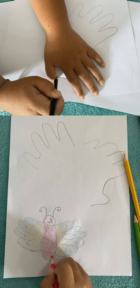 An 8-year-old child coloring a smiling, front-facing handprint butterfly drawing with pink and yellow colored pencils.
