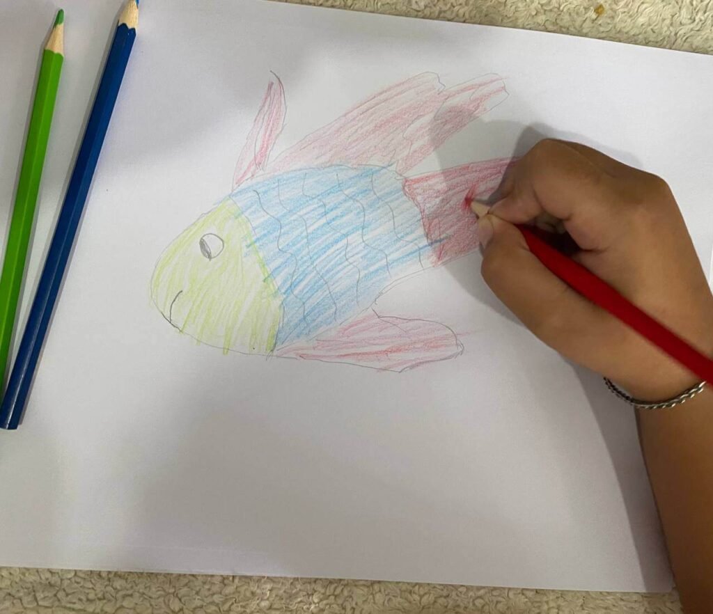 A child coloring a hand-traced fish drawing that has a yellow head, a blue body with wavy scales, and red fins.