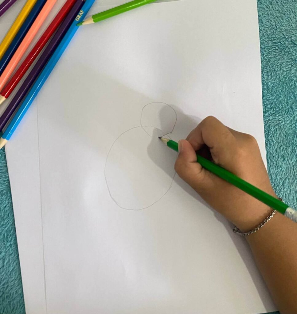 A child's hand holding a green pencil, sketching an oval and a small circle on white paper to form a bunny's body without a neck.