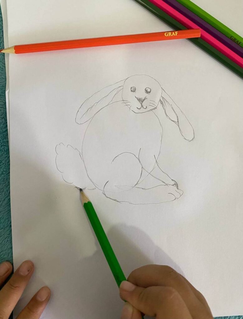 A close-up of a child using a pencil to sketch a fluffy tail on a bunny drawing, which also features a Y-shaped nose and whiskers.