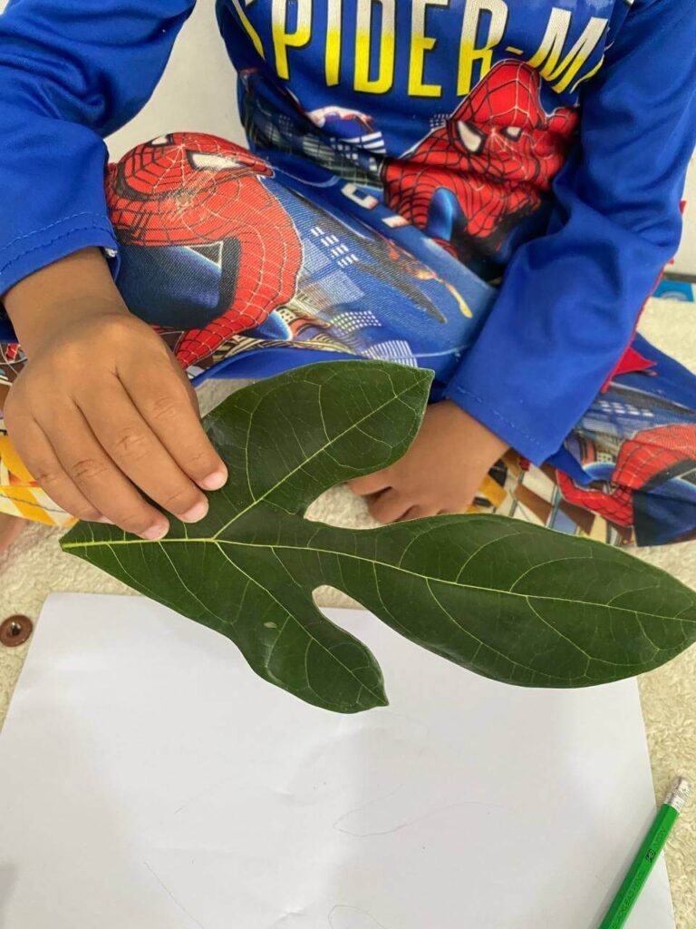 A close-up of a child wearing a blue Spiderman shirt, holding a green jackfruit leaf over a blank sheet of white paper.